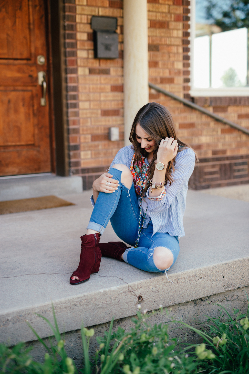 Girl sitting at front door with blue stripe jacket with pom pom tassels distressed jeans and maroon wedges