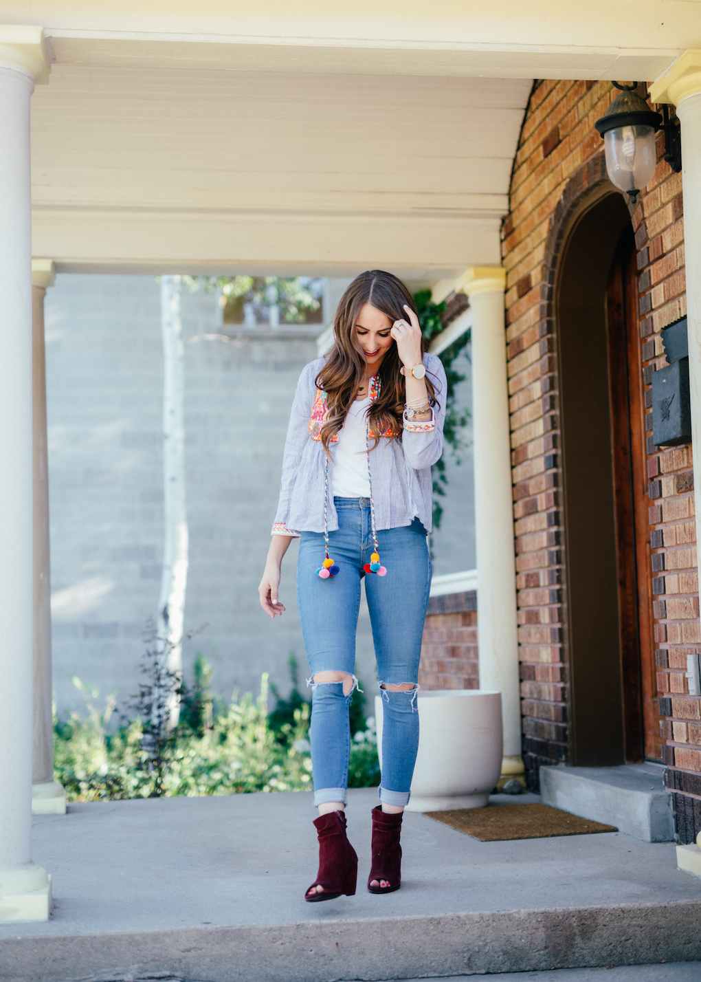 Girl standing at front door with blue stripe jacket with pom pom tassels distressed jeans and maroon wedges
