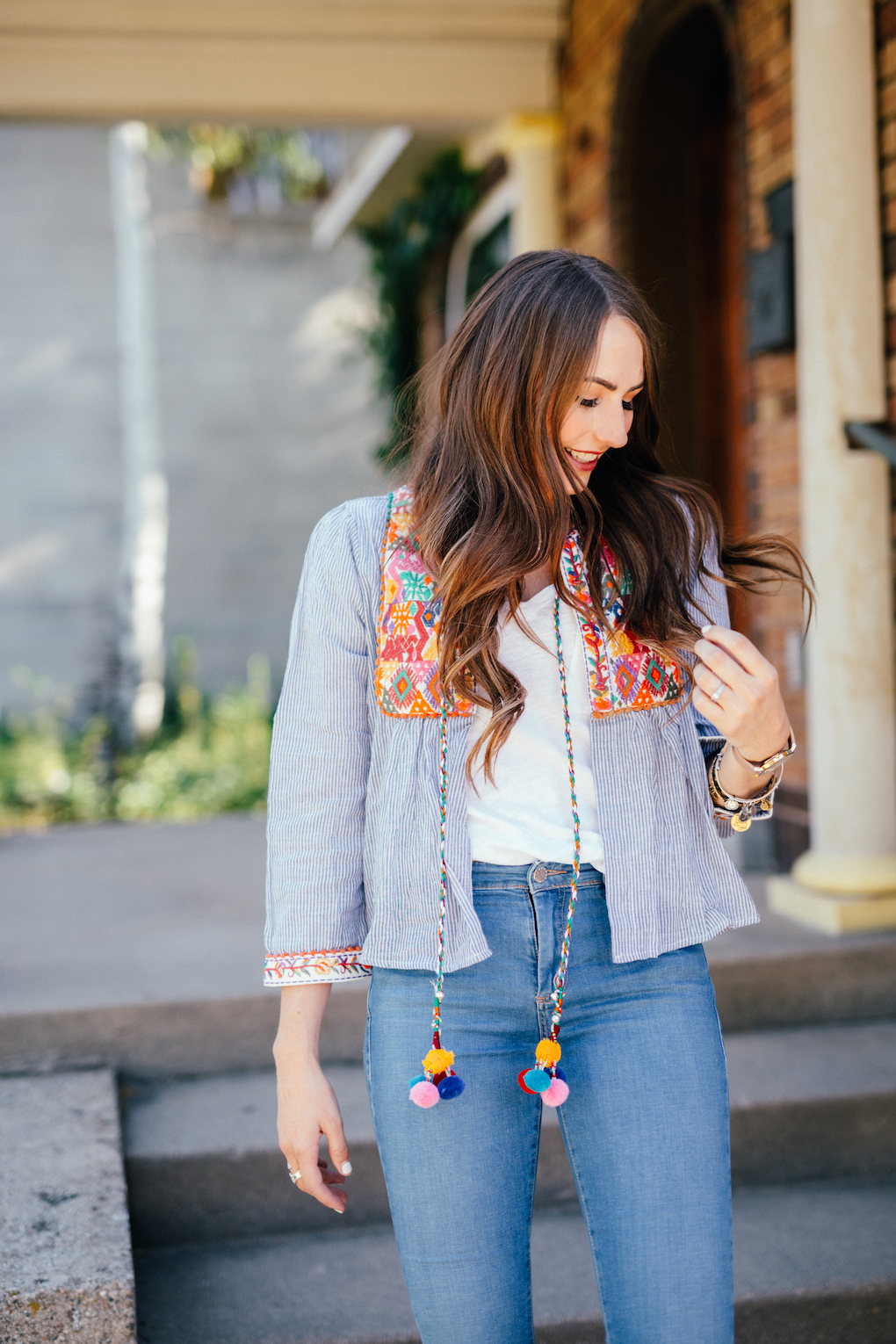 Girl with loose brown hair wearing distressed denim jeans and blue stripe embroidered jacket with pom pom tassels
