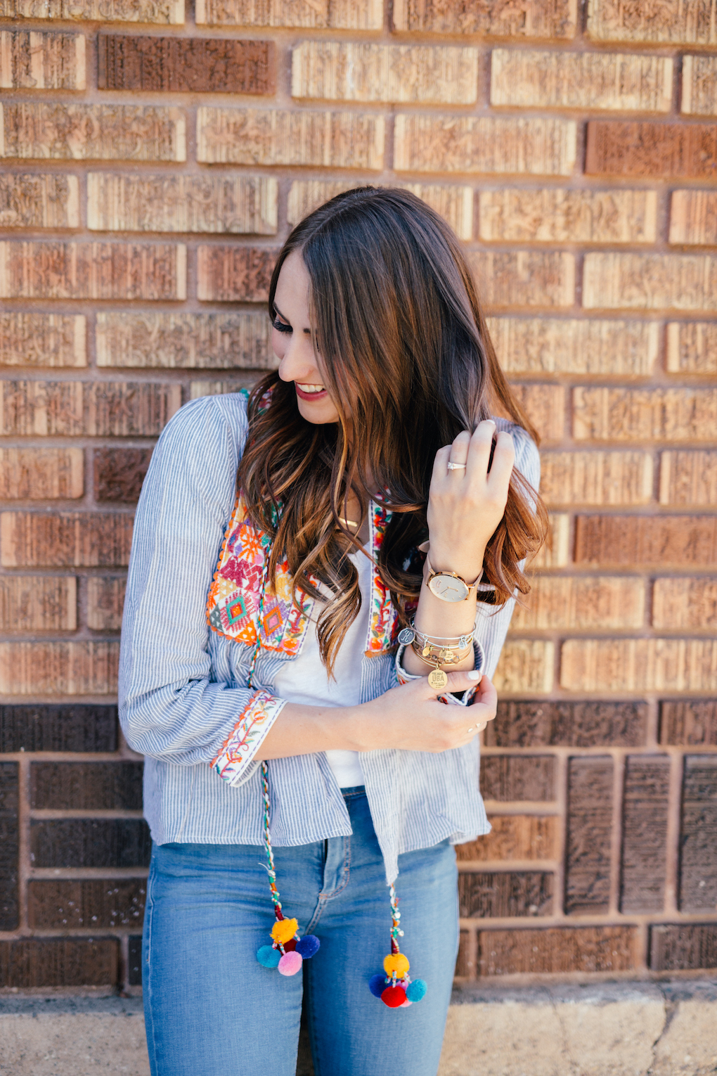 Girl with loose brown hair wearing distressed denim jeans and blue stripe embroidered jacket with pom pom tassels