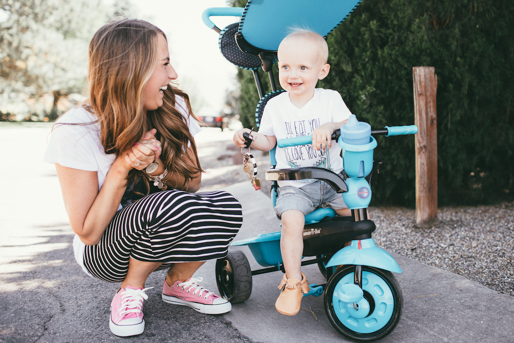 blonde little boy in bright blue SmarTrike with blue cup wearing white v neck tee cut off shorts and moccs and mom next to him wearing black stripe skirt