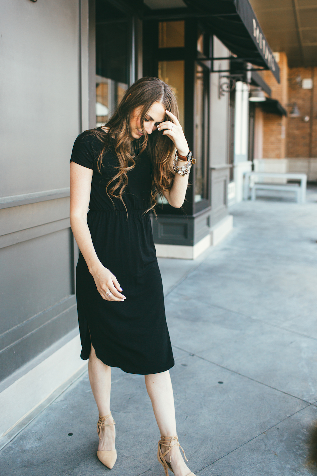 Girl with long brown hair curled wearing black dressed with rouched waist band and nude heels that tie around the ankle with arvo watch and alex and ani bracelets stacked