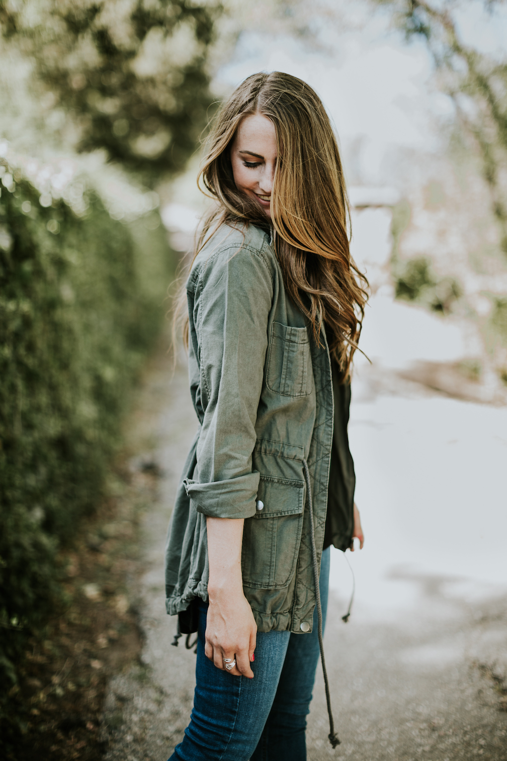 girl looking down with eyes closed wearing green army jacket with the sleeves rolled and loose curled brown hair
