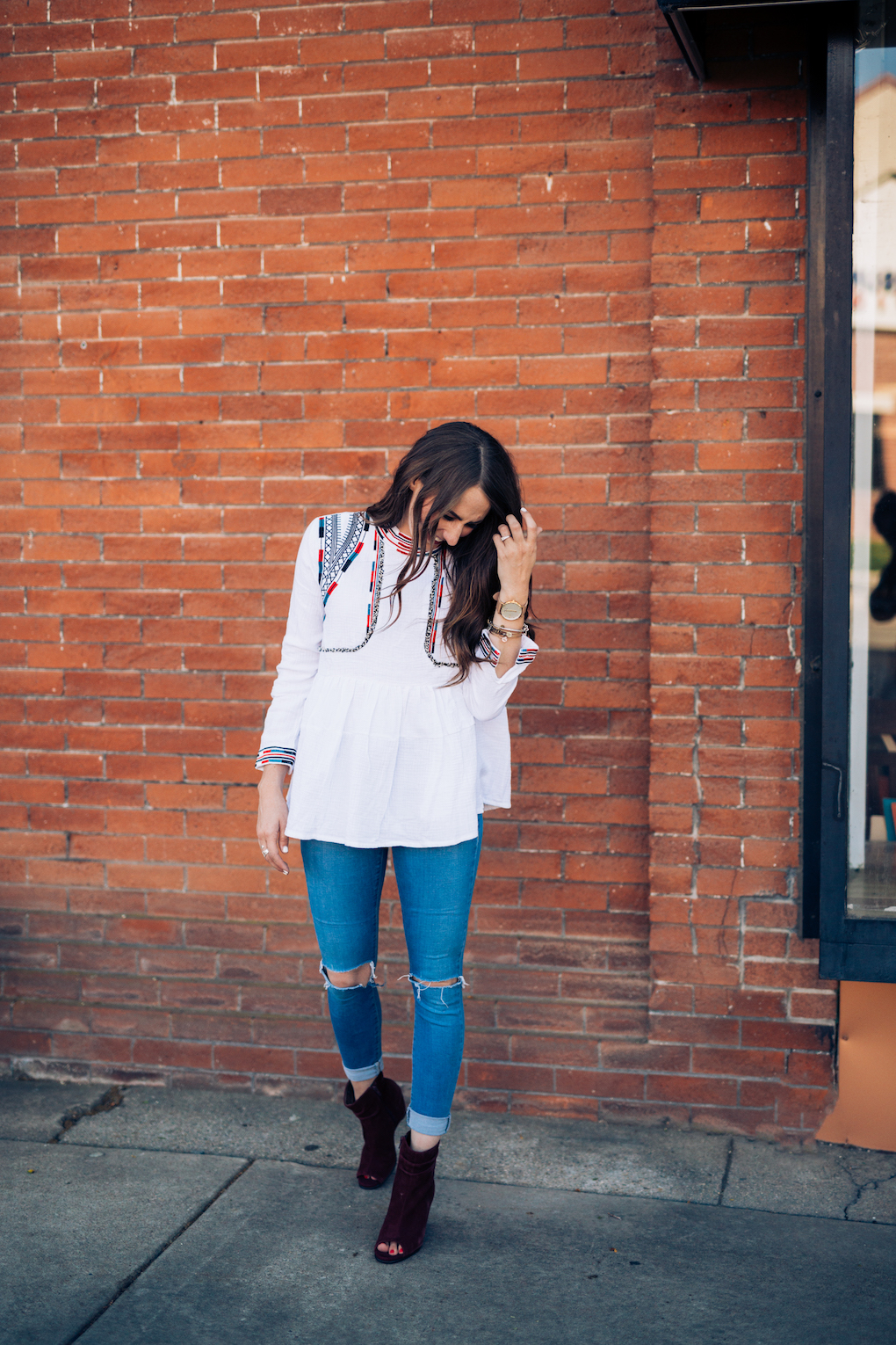 Girl in white peplum top with bright colored embroidery and skinny jeans with maroon booties in front of red wall