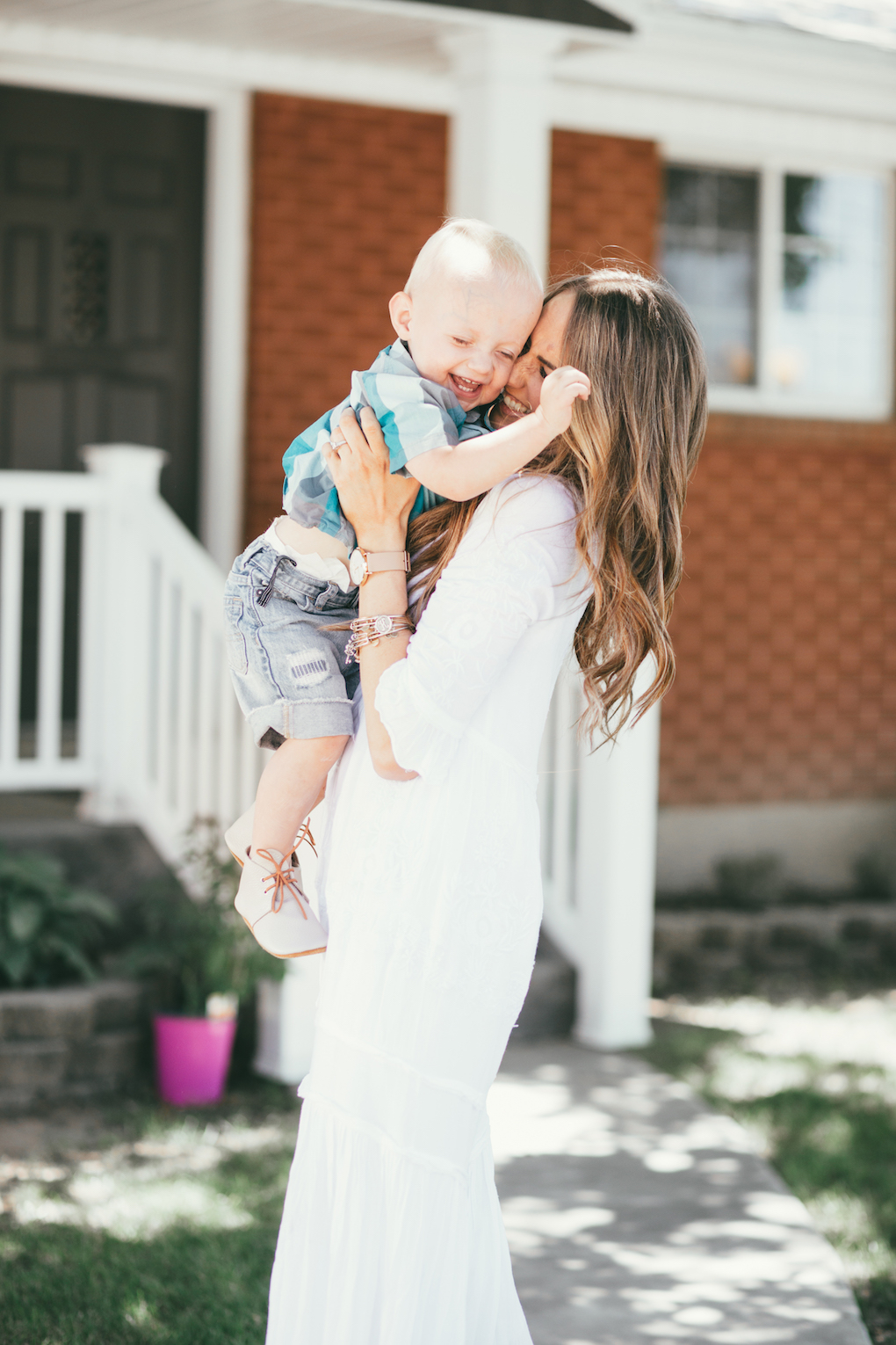 mom and little boy standing in front of red brick house mom wearing white maxi dress with long brown hair loosely curled and blonde highlights little boy wearing turquoise button up with cut off shorts and mon petite shoes