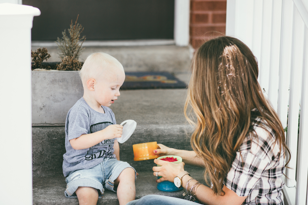 Little Boy in Grey Tee Shirt wearing denim cutoff shorts and mon petite shoes playing with zoli snack container with his mom wearing a plaid short sleeved top and brown loose curls