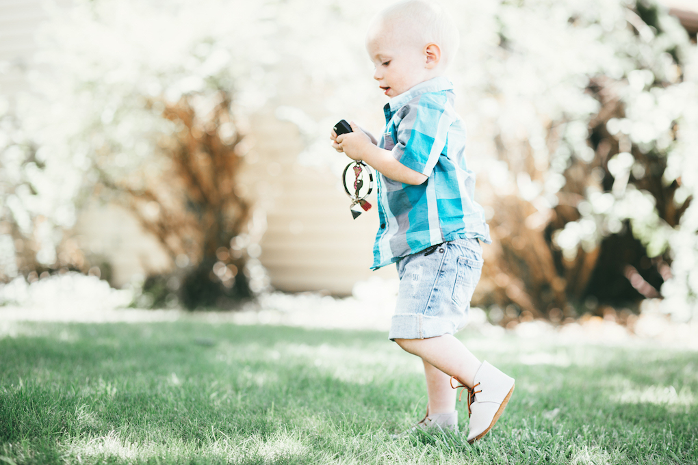 blonde little boy playing on the grass with keys wearing a checked turquoise button up shirt, cut off shorts, and mon petite shoes