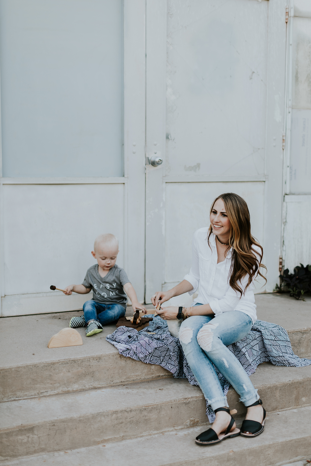 mom and little boy playing on stairs with captain silly pants swaddle blanket wooden toys wooden car mom wearing white button up top with skinny distressed jeans and pons little boy in grey graphic tee skinny distressed denim and freshly picked moccasins