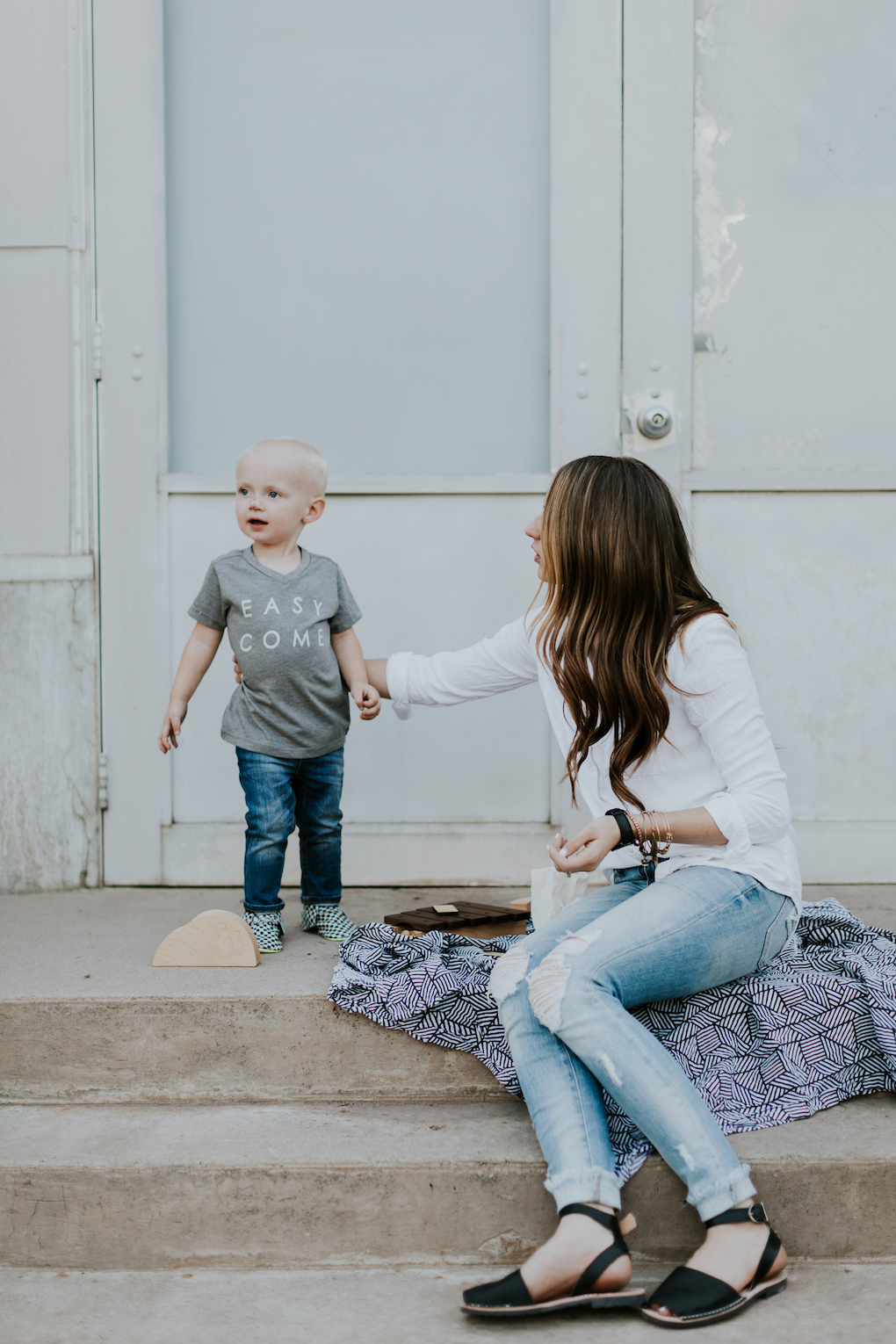 mom and little boy playing on stairs with captain silly pants swaddle blanket wooden toys wooden car mom wearing white button up top with skinny distressed jeans and pons little boy in grey graphic tee skinny distressed denim and freshly picked moccasins