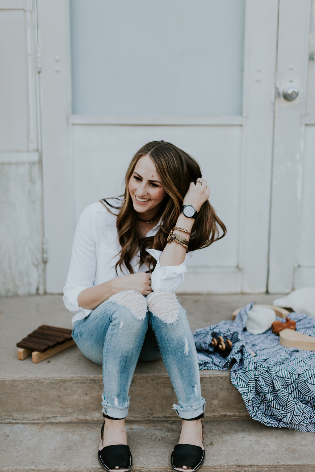 mom sitting on steps with captain silly pants swaddle blanket and wooden toys wearing white button up shirt arvo black watch alex and ani bracelets with choker on and distressed denim with pons shoes