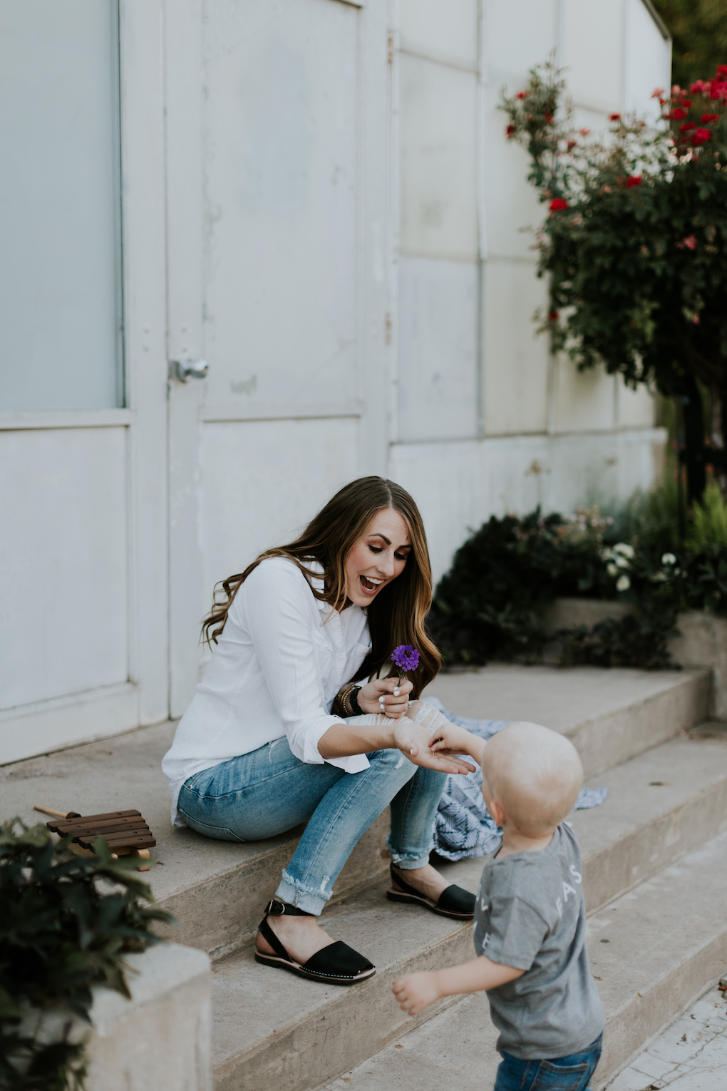 mom sitting on steps with captain silly pants swaddle blanket and wooden toys wearing white button up shirt arvo black watch alex and ani bracelets with choker on and distressed denim with pons shoes