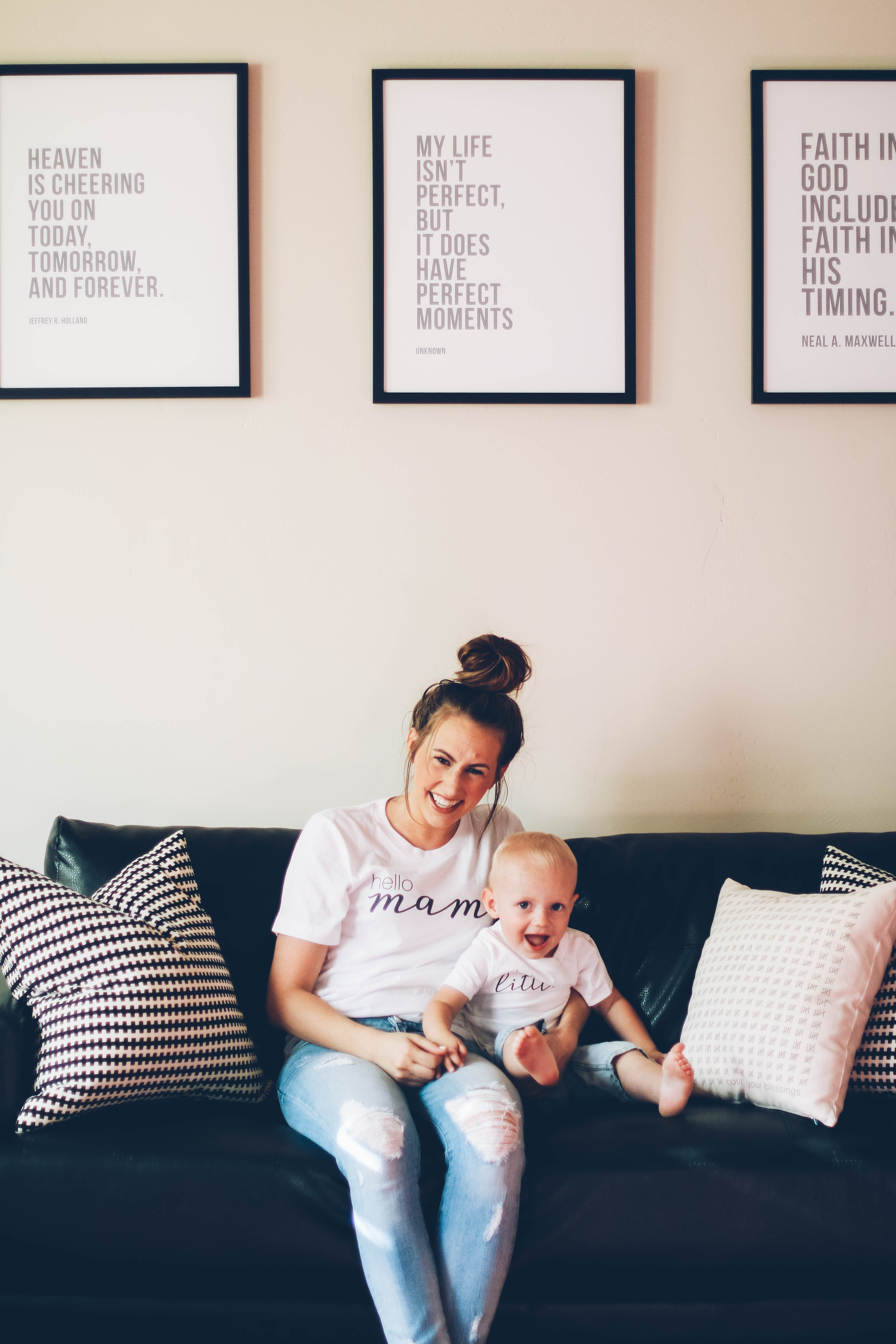 Mom and little boy sitting on modern black leather couch with lds framed prints on wall and ruby claire white tee shirts on with denim shorts and denim skinny jeans