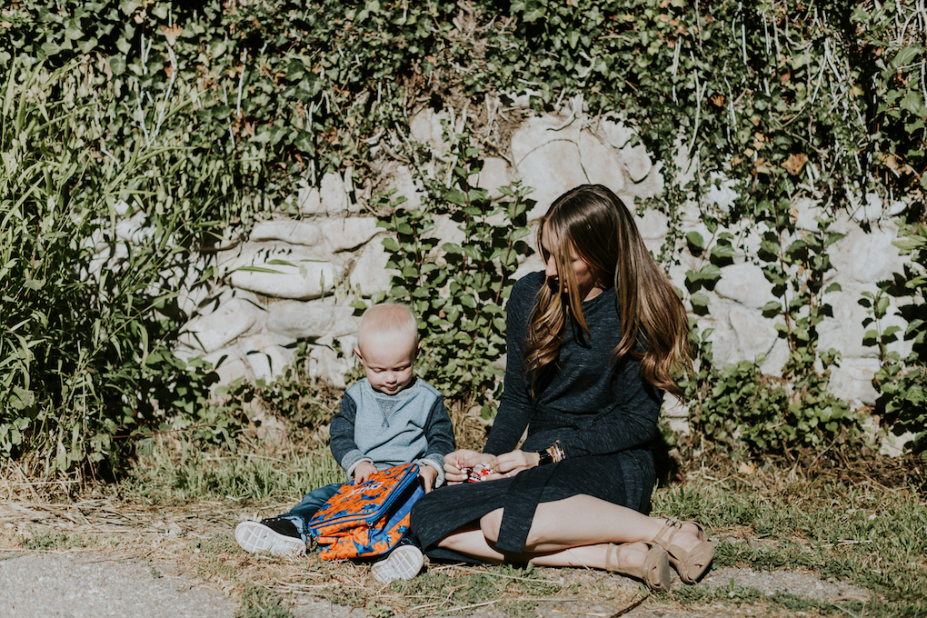 mom in navy tie dress little boy in crew neck sweatshirt with denim joggers on sitting on the cement with vines behind them and a dinosaur lunch box