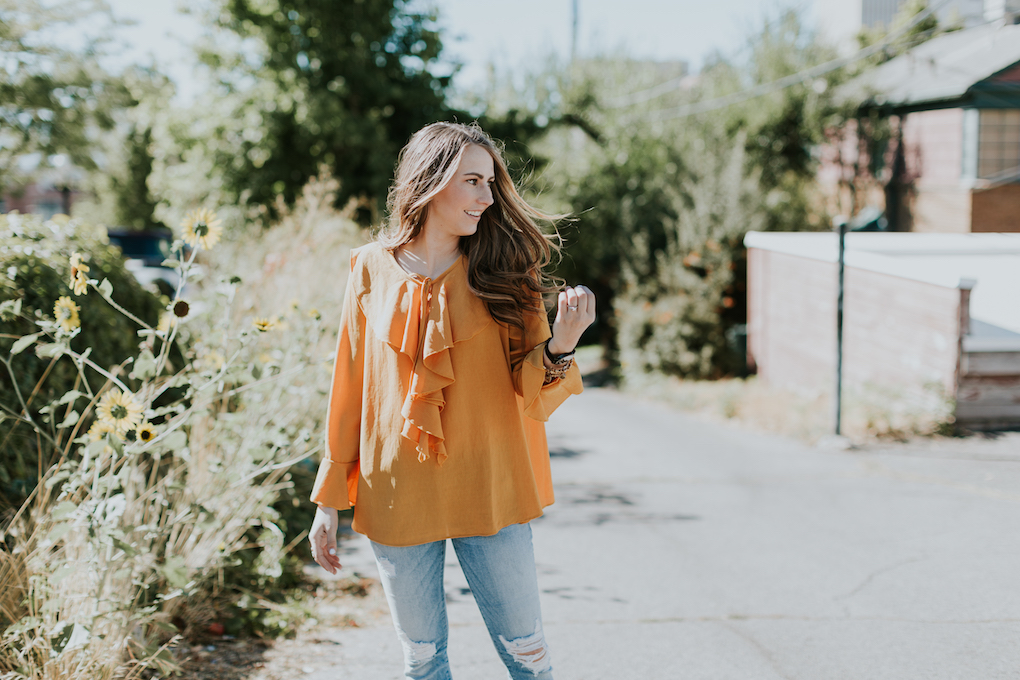 girl with brown caramel hair wearing mustard orange sheer ruffle top with distressed jeans