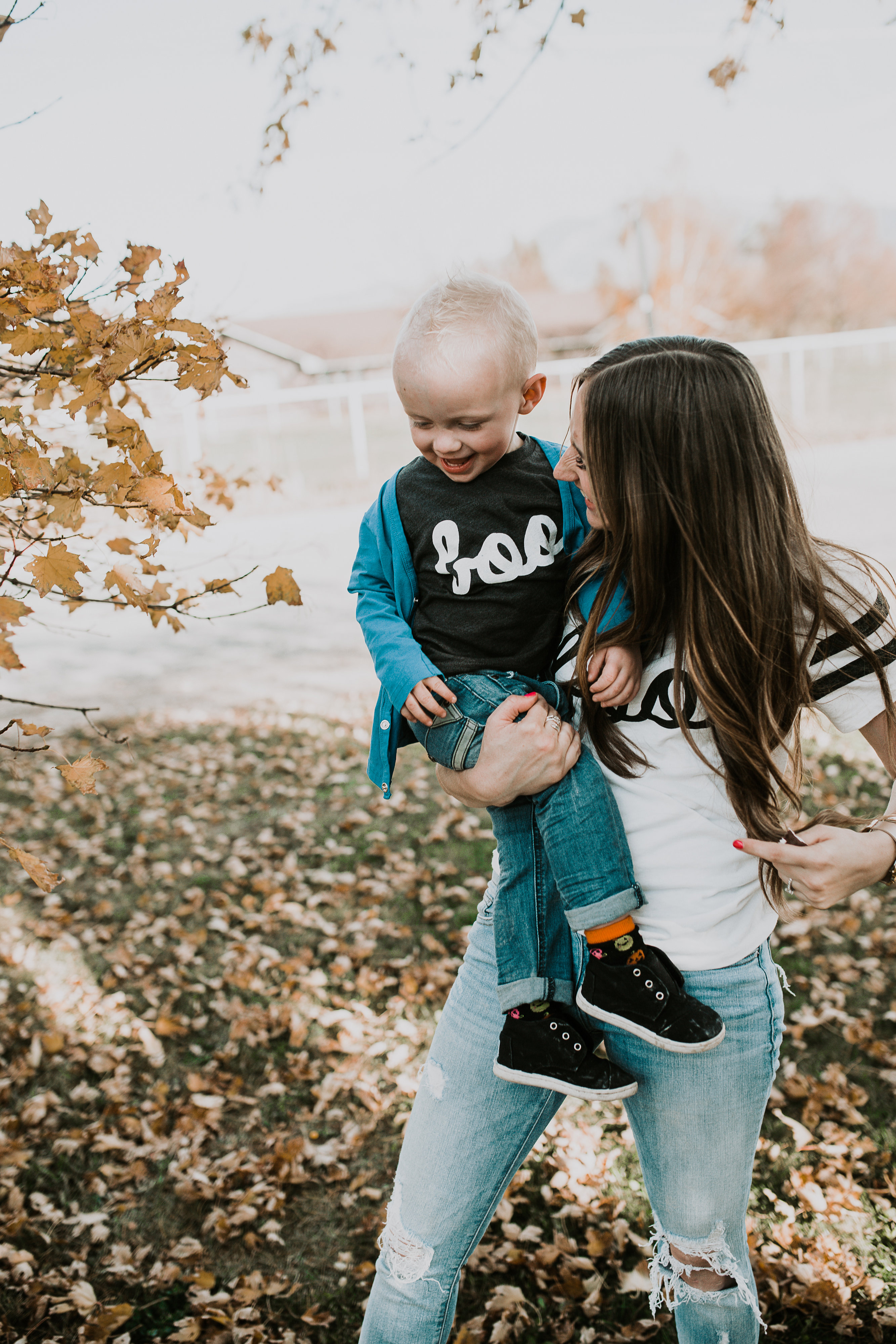 mom and little boy in BOO halloween tees playing in the leaves mom has light brown loosely curled hair and wearing distressed denim little boy in june and january cardigan