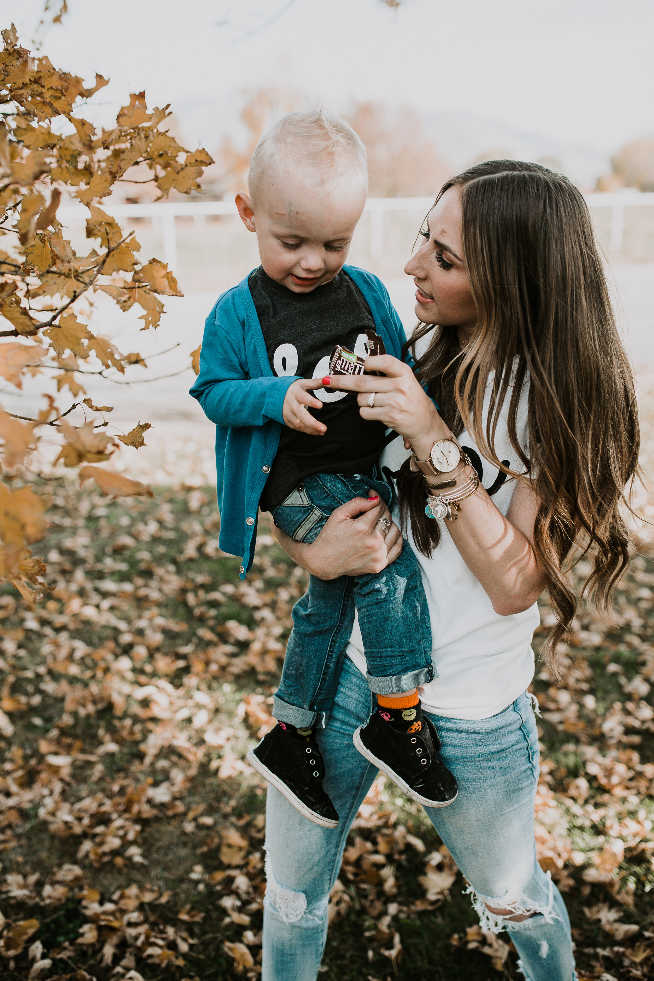 mom and little boy in BOO halloween tees playing in the leaves mom has light brown loosely curled hair and wearing distressed denim little boy in june and january cardigan