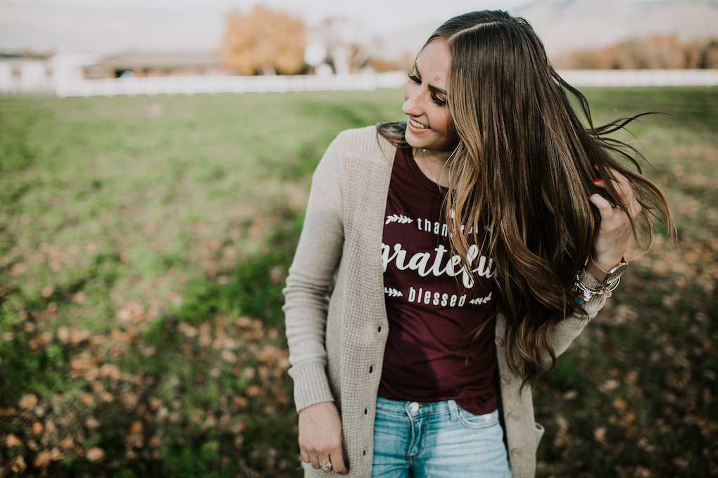 girl standing in field in wine colored grateful tee a long cream cardigan and distressed denim jeans with loosely curled brown hair