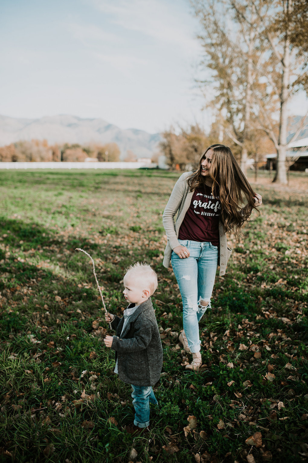 mom and little boy playing in field little boy in tweed jacket and plaid button up shirt with skinny jeans and brown boots mom in long cream cardigan a wine colored grateful tee and distressed denim