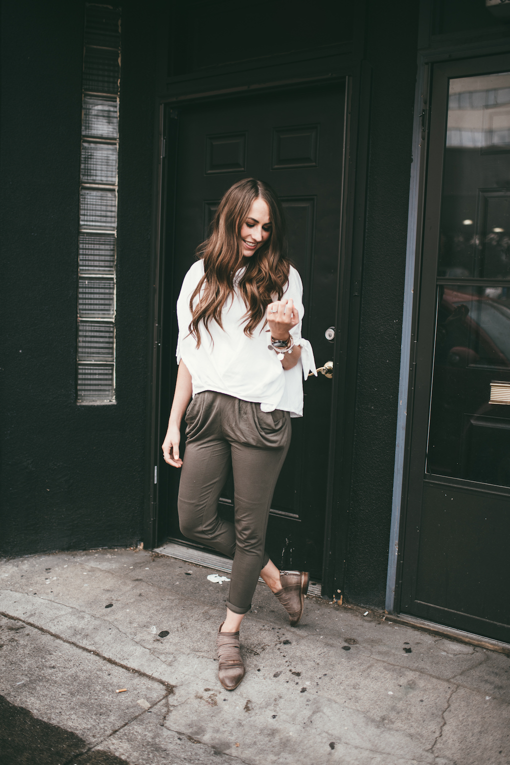 olive green knit green pants with oversized white top on girl standing in front of all black doorway with long brown curled hair
