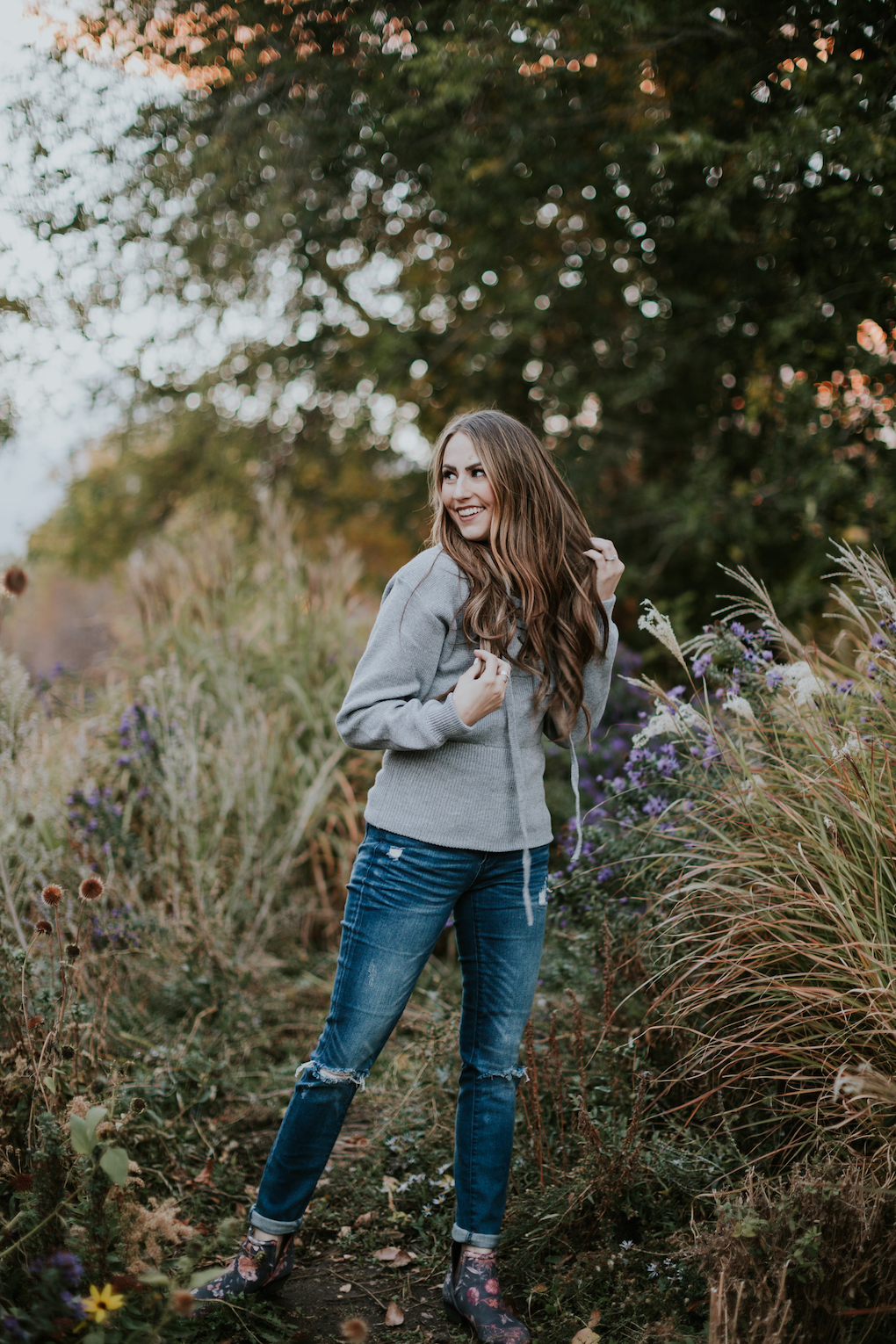 girl in grey lace sweater and distressed jeans standing in greenery with long caramel brown hair loosely curled