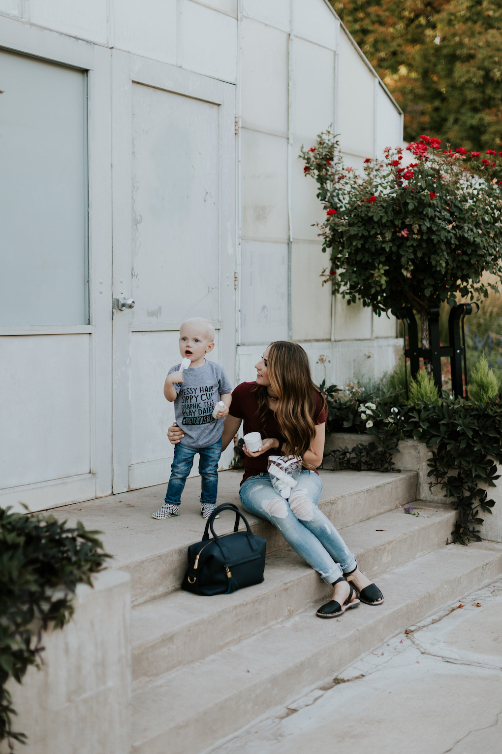 mom and little boy sitting on the stairs with baby pibu products little boy in graphic tee and jeans mom in distressed denim a maroon top and a navy handbag