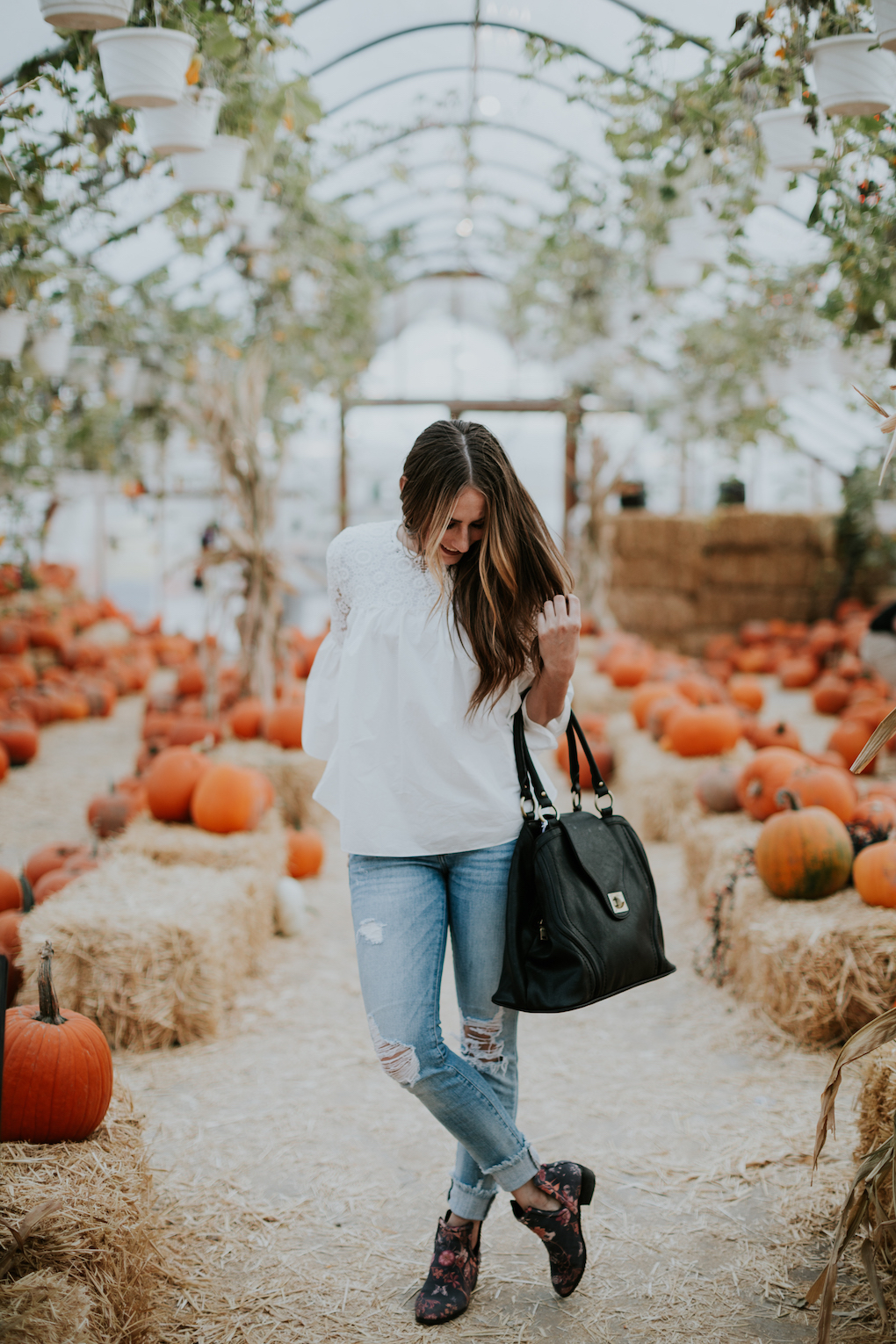 girl wearing white lace top with distressed denim and brown booties standing in a pumpkin patch with black diaper bag
