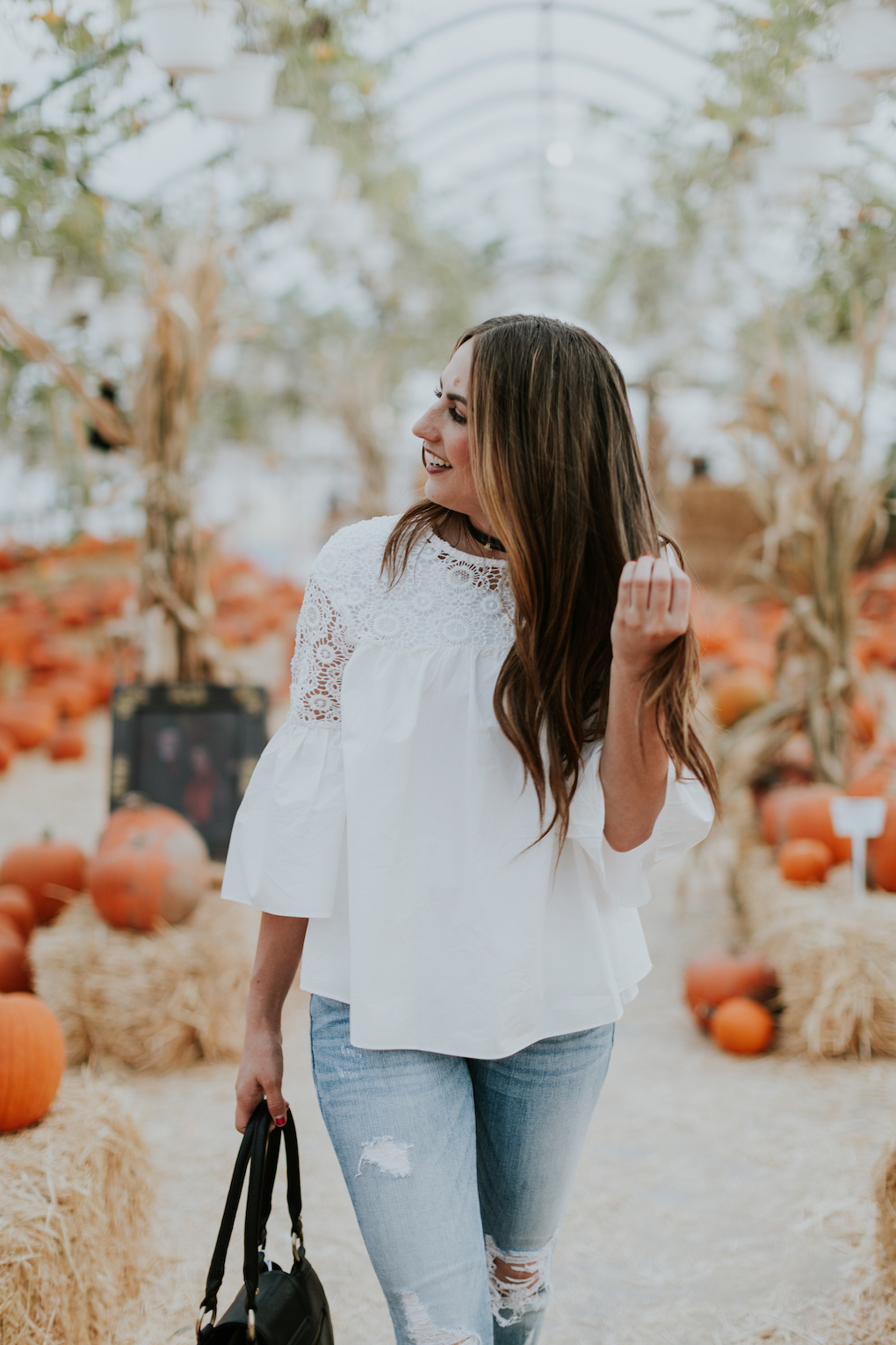 girl wearing white lace top with distressed denim and brown booties standing in a pumpkin patch with black diaper bag