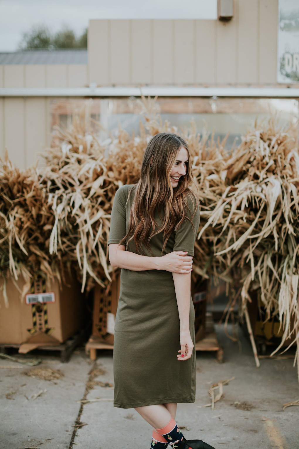 girl standing in front of cornstalks wearing a green midi dress, tall floral socks, strap black booties, and a choker with long brown caramel hair loosely curled