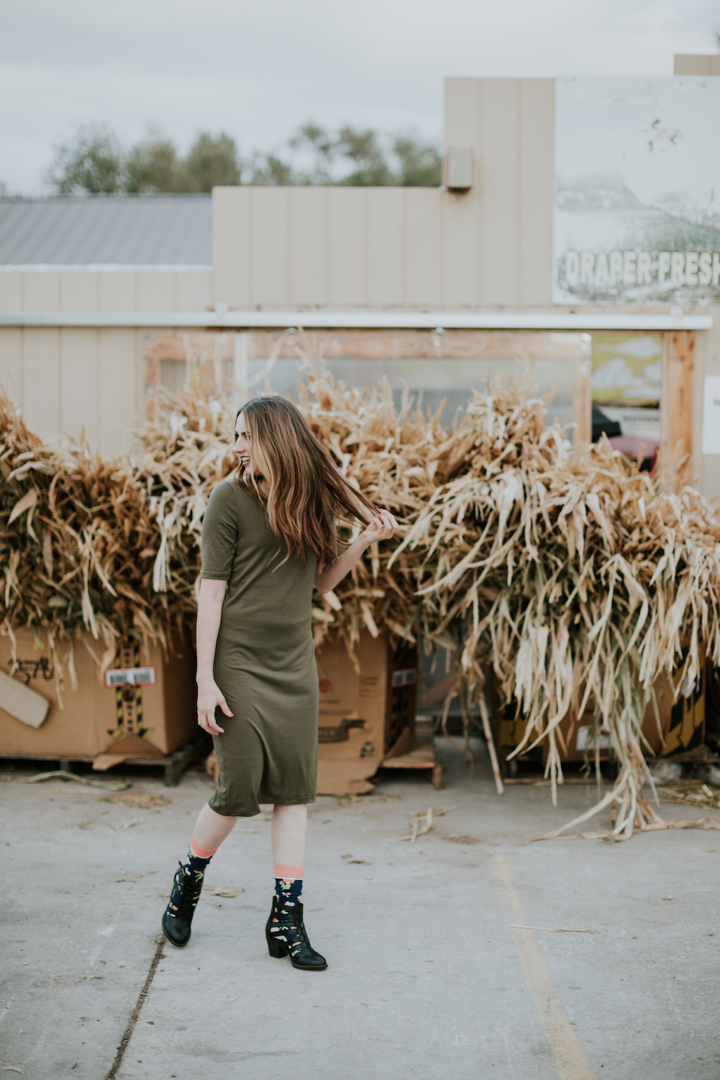 girl standing in front of cornstalks wearing a green midi dress, tall floral socks, strap black booties, and a choker with long brown caramel hair loosely curled