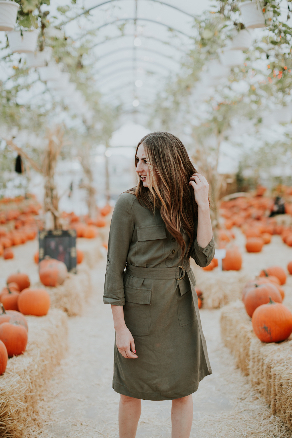 Girl standing in green house pumpkin patch with a green military style midi dress on with long brown caramel hair in loose curls and black cut out booties on