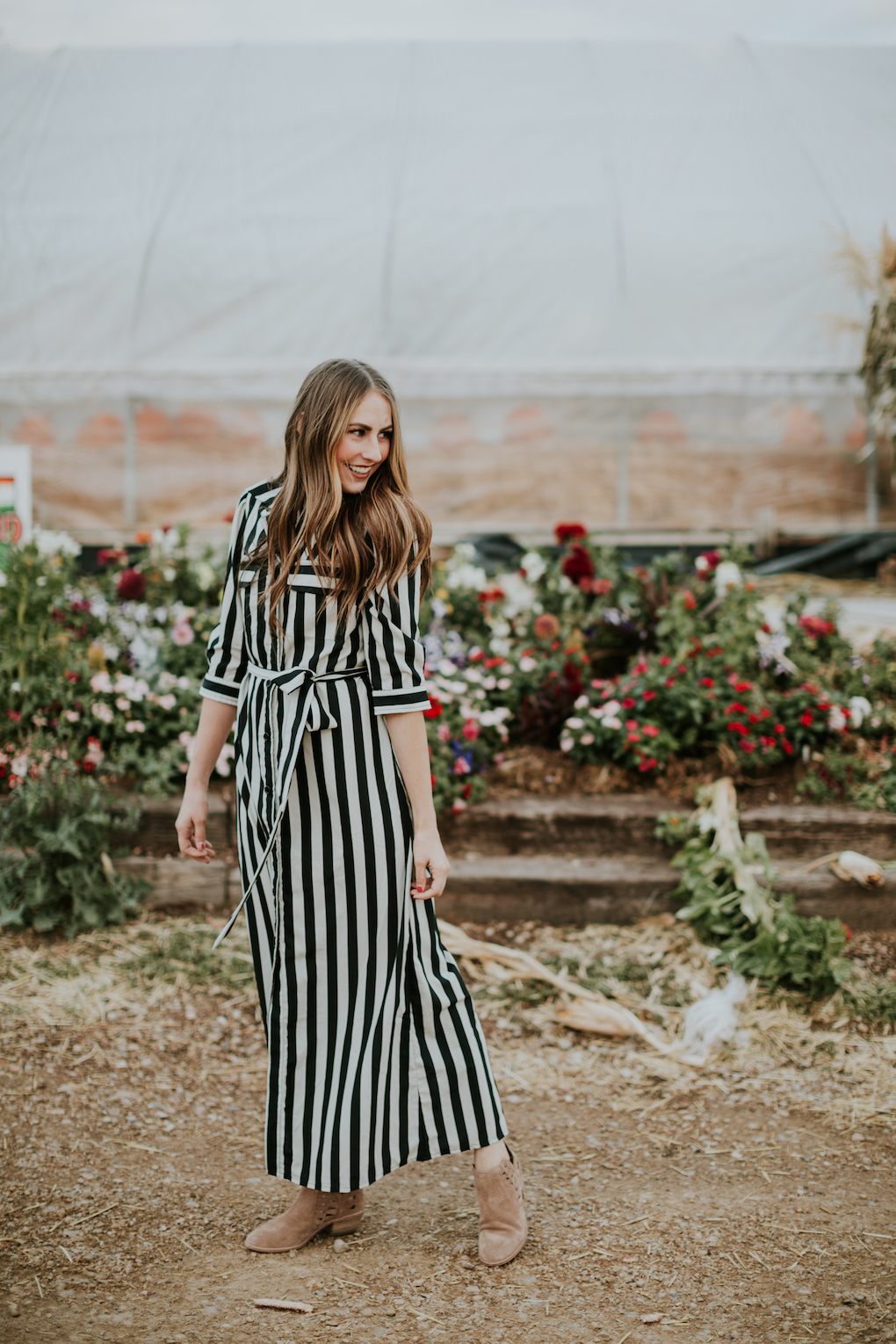 girl standing in front of green house pumpkin patch wearing a black and white stripe long maxi dress with loosely curled brown hair with caramel highlights
