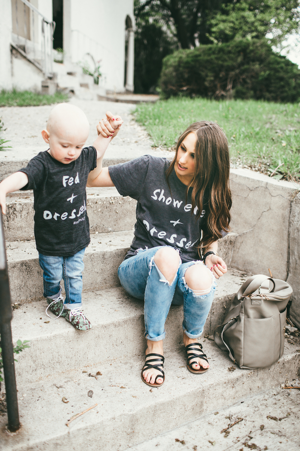 Mom and little boy in matching graphic tees, distressed denim sitting on cement stairs with a Fawn Design bag in Grey