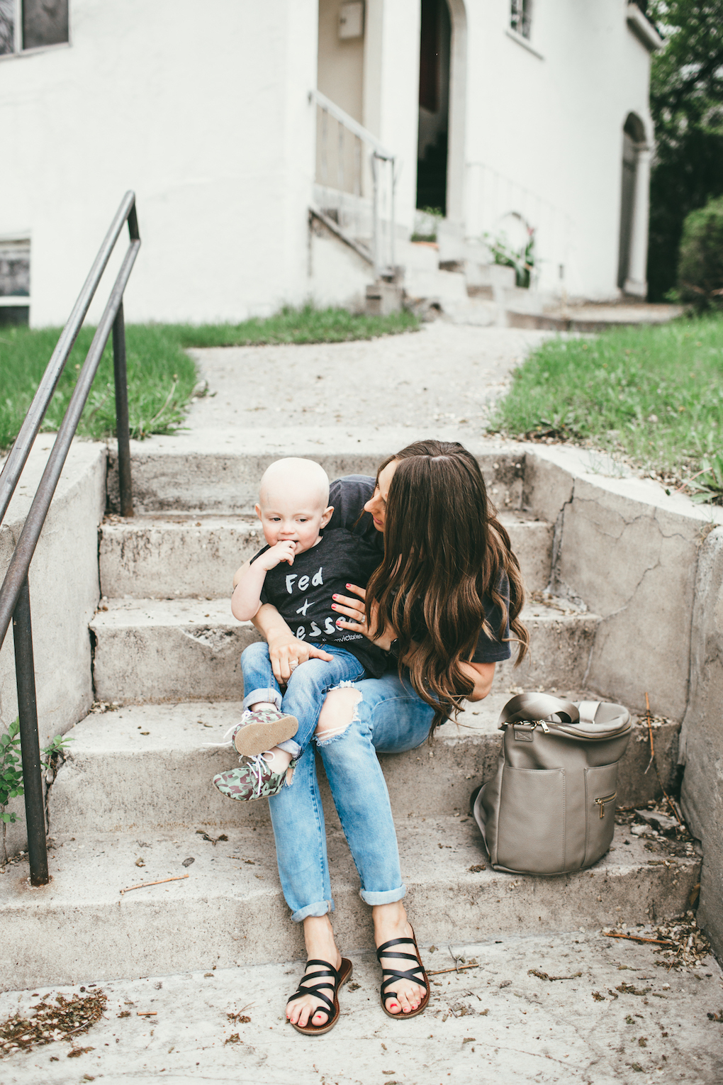 Mom and little boy in matching graphic tees, distressed denim sitting on cement stairs with a Fawn Design bag in Grey