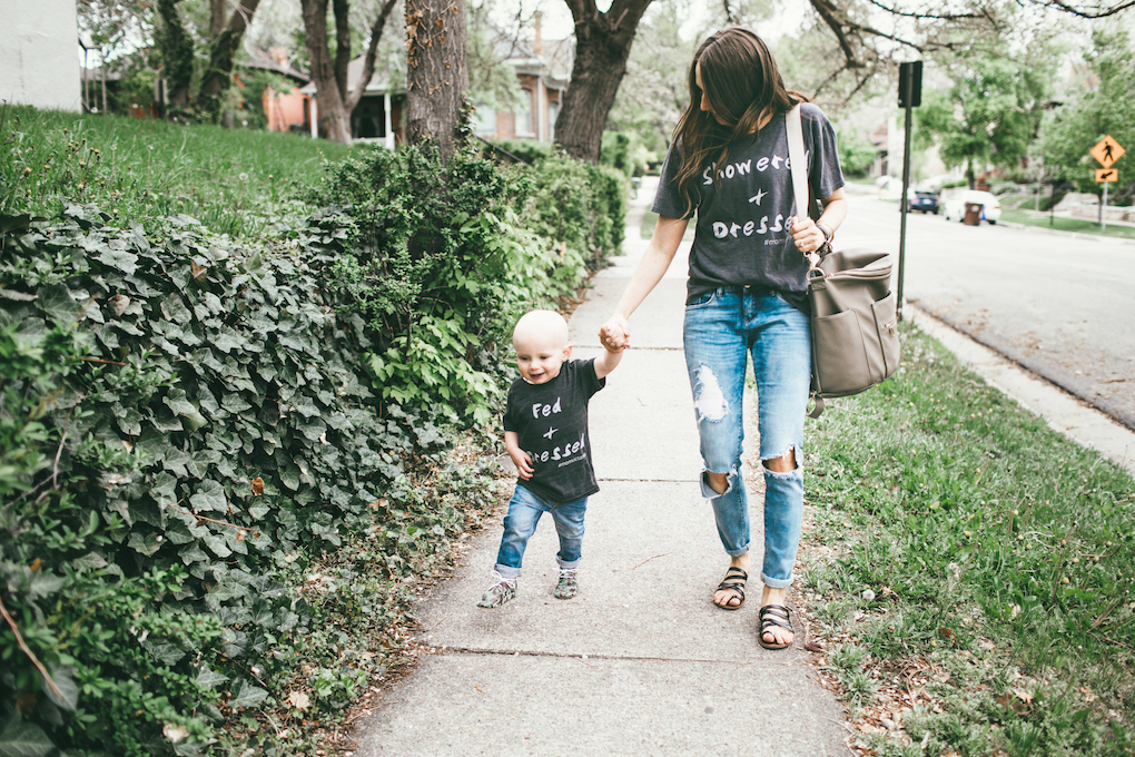 Mom and little boy in matching graphic tees, distressed denim walking down sidewalk with a Fawn Design bag in Grey