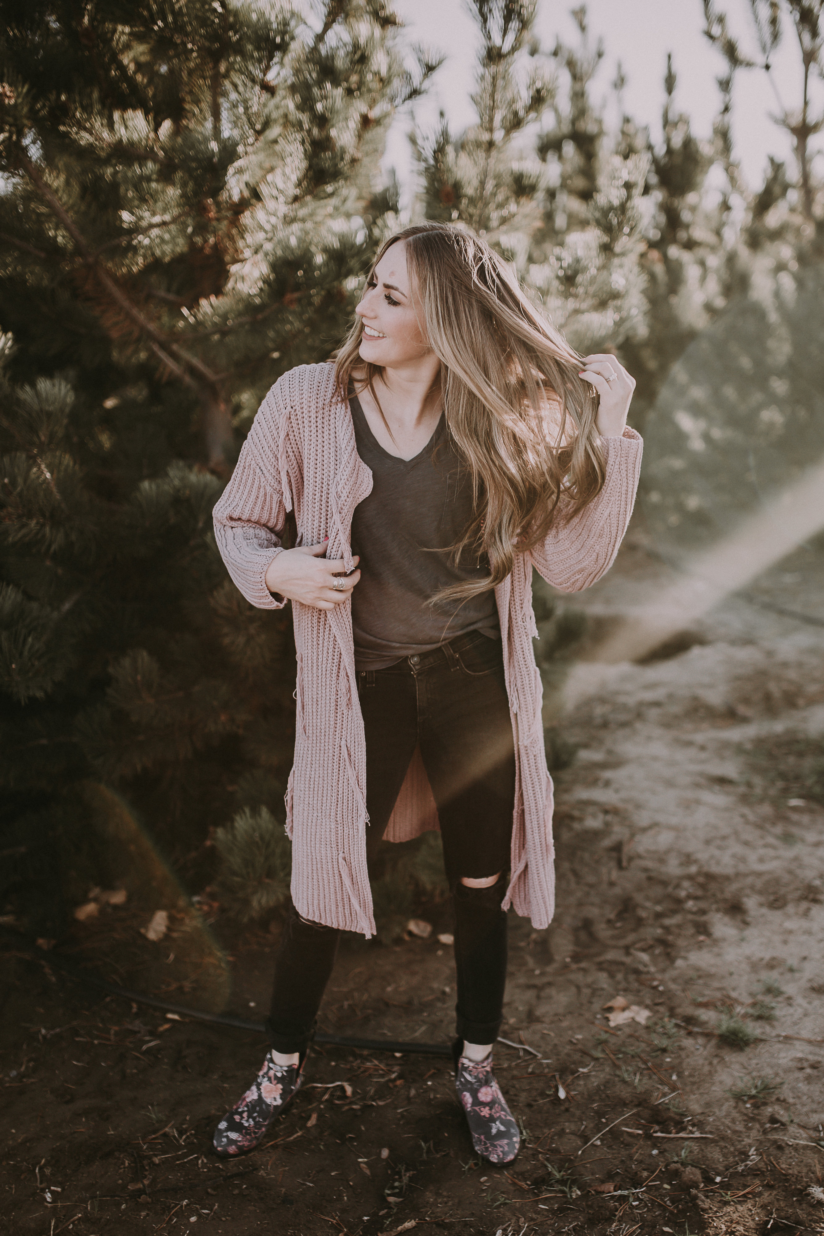 girl in light pink long thick cardigan with grey tee shirt floral booties and black distressed jeans with long loose curled brown and caramel long hair