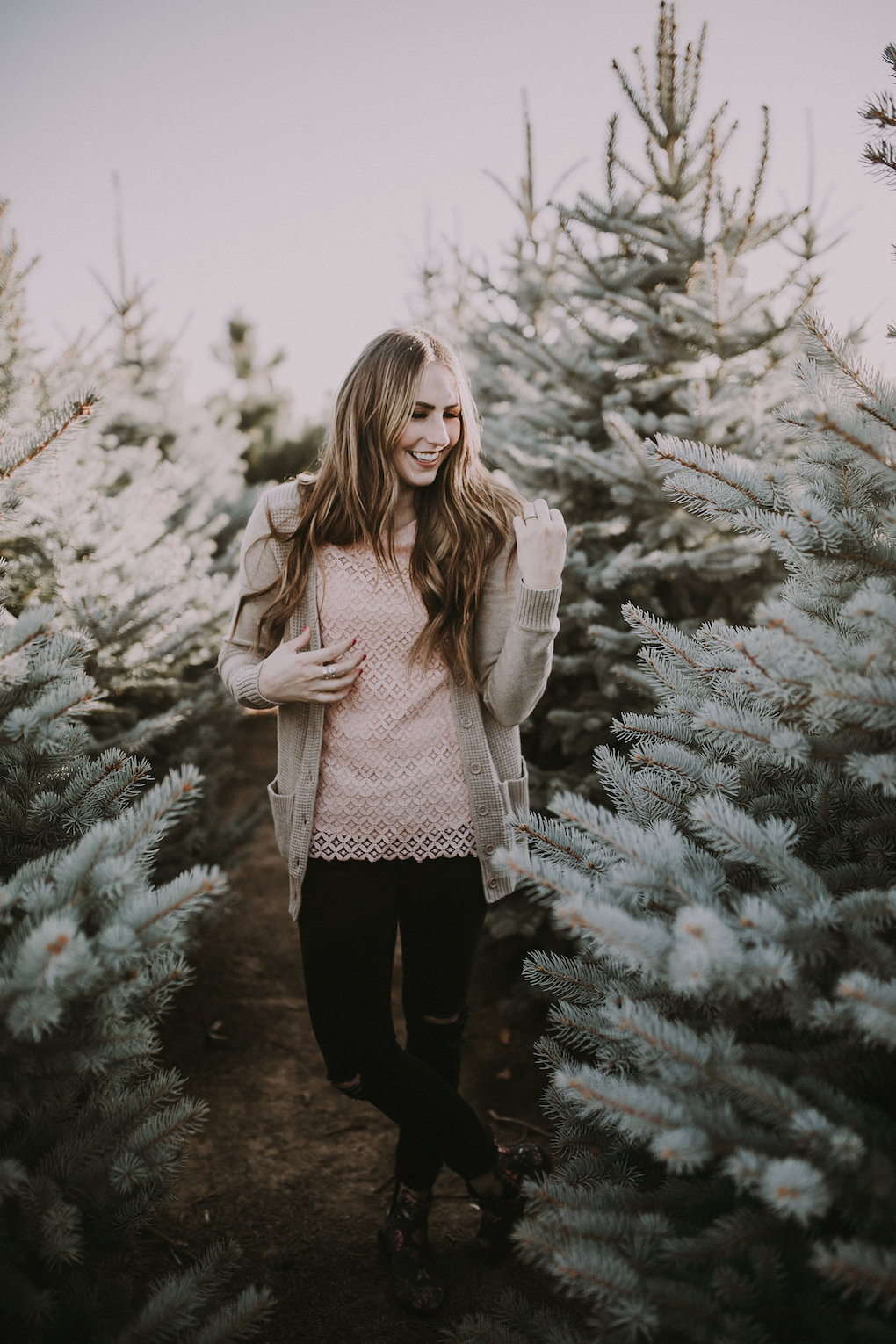 girl standing in Christmas Tree farm with long loosely curled brown hair with caramel highlights wearing downeast lace pink top with knitted thick cream cardigan and black jeggings