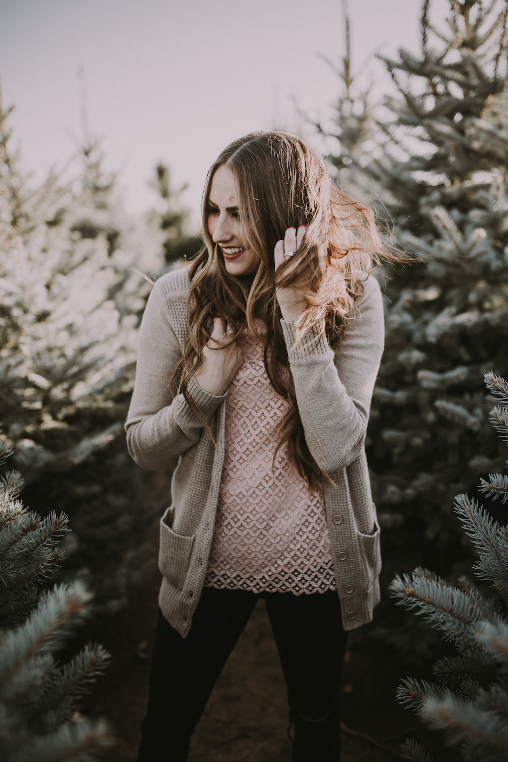 girl standing in Christmas Tree farm with long loosely curled brown hair with caramel highlights wearing downeast lace pink top with knitted thick cream cardigan and black jeggings