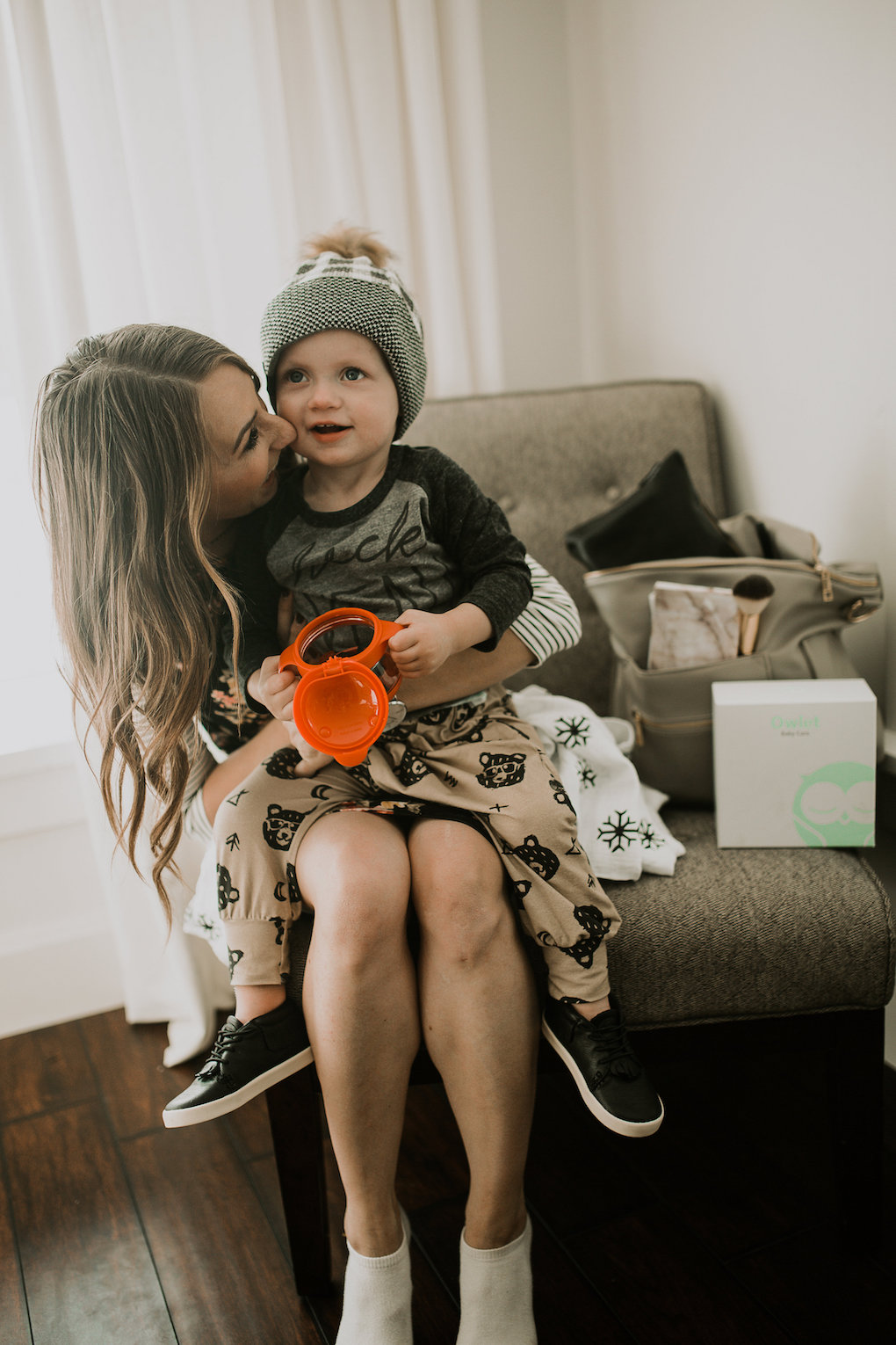 mom and little boy sitting on chair with fawn design diaper bag and diaper clutch owlet baby monitor little adi co clothes and hat ruby claire dress and ubbi snack container