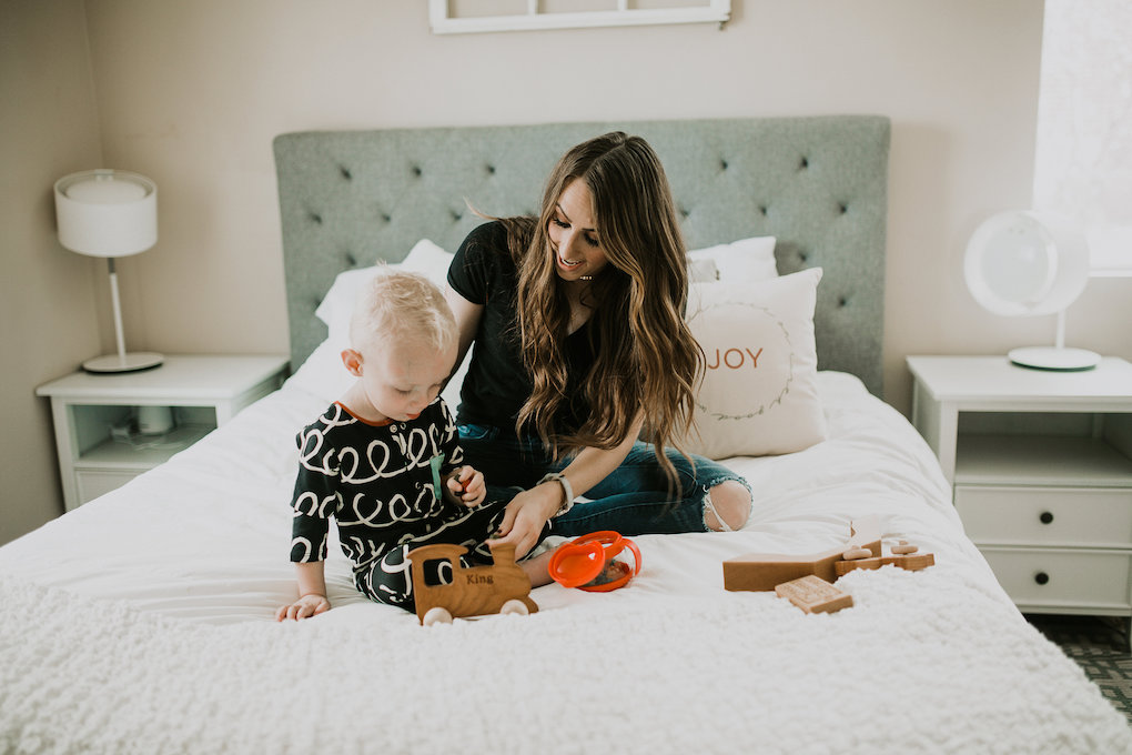 mom and little boy playing on the bed with little boy in rags to raches romper playing with bannor toys and latterday home pillow on the bed