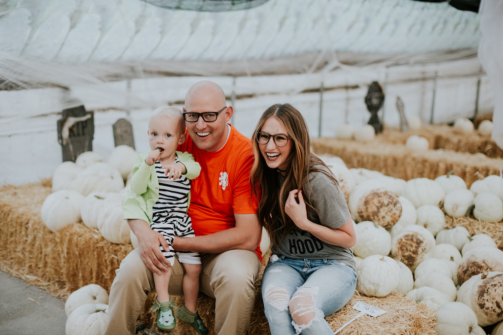 family sitting and laughing at the pumpkin patch mom in grey tee and glasses with distressed jeans dad in orange tee and khaki pants little boy in black and white romper and green cardigan