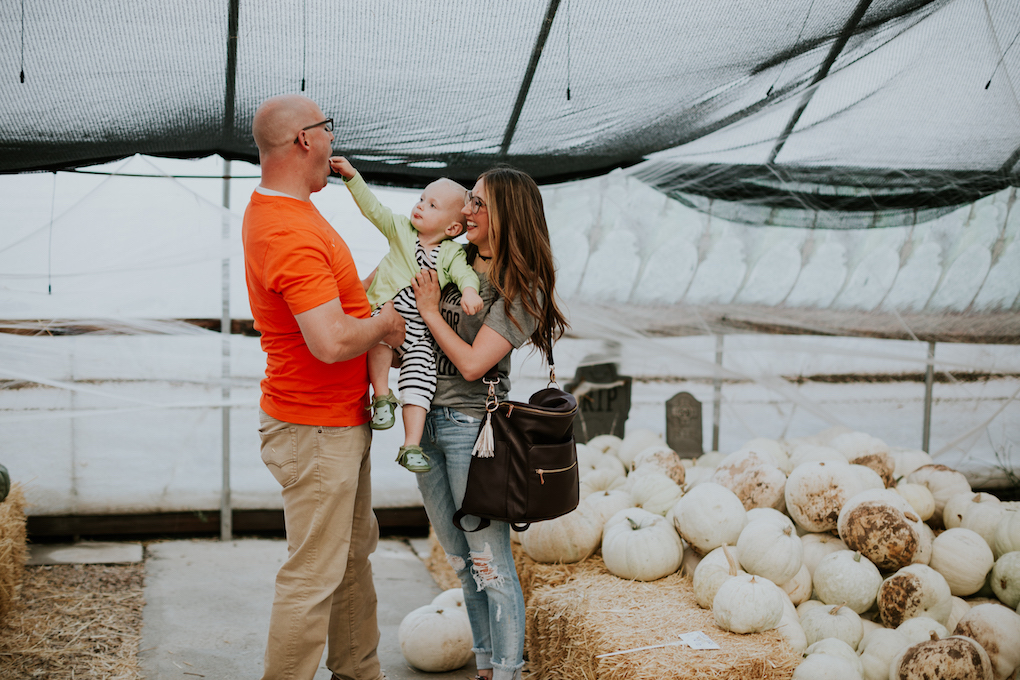 family sitting and laughing at the pumpkin patch mom in grey tee and glasses with distressed jeans dad in orange tee and khaki pants little boy in black and white romper and green cardigan