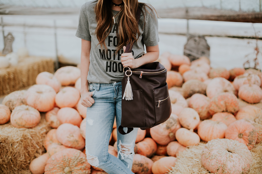 girl standing in pumpkin patch with grey graphic tee distressed jeans and plum fawn design bag