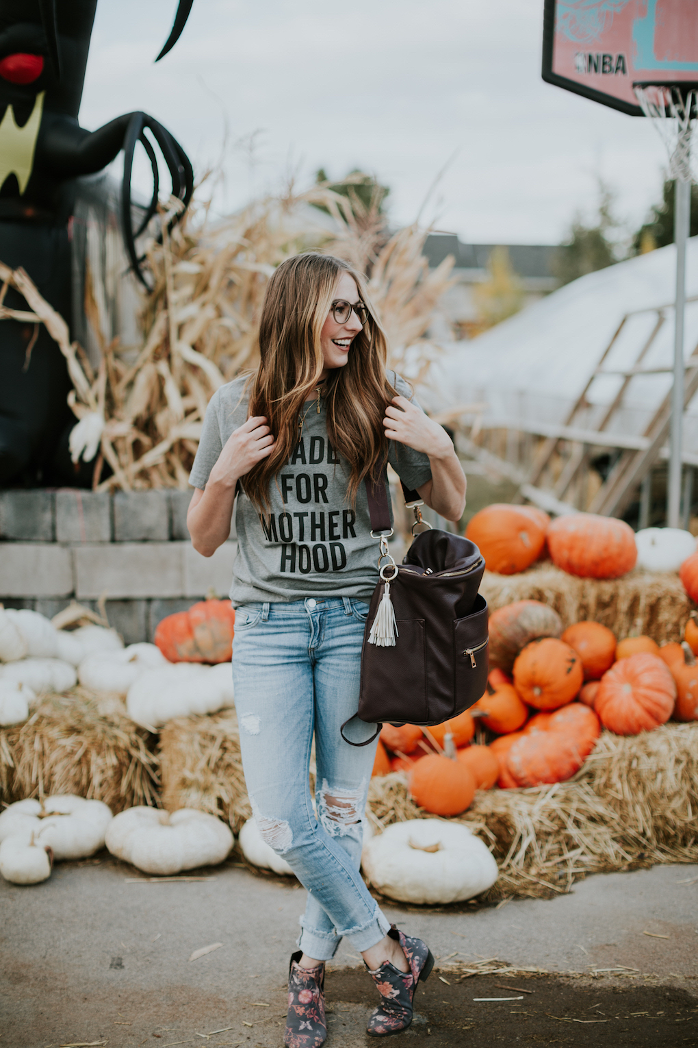 girl standing in pumpkin patch with grey graphic tee distressed jeans and plum fawn design bag