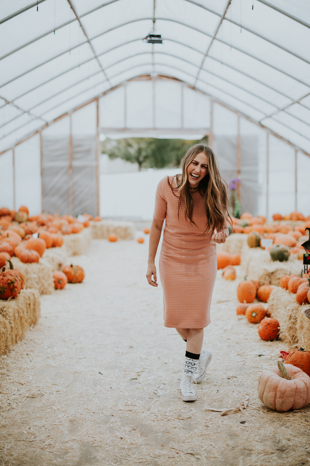 girl in quilted pink dress with tall glasses socks and converse on standing at pumpkin patch with loosely curled brown hair