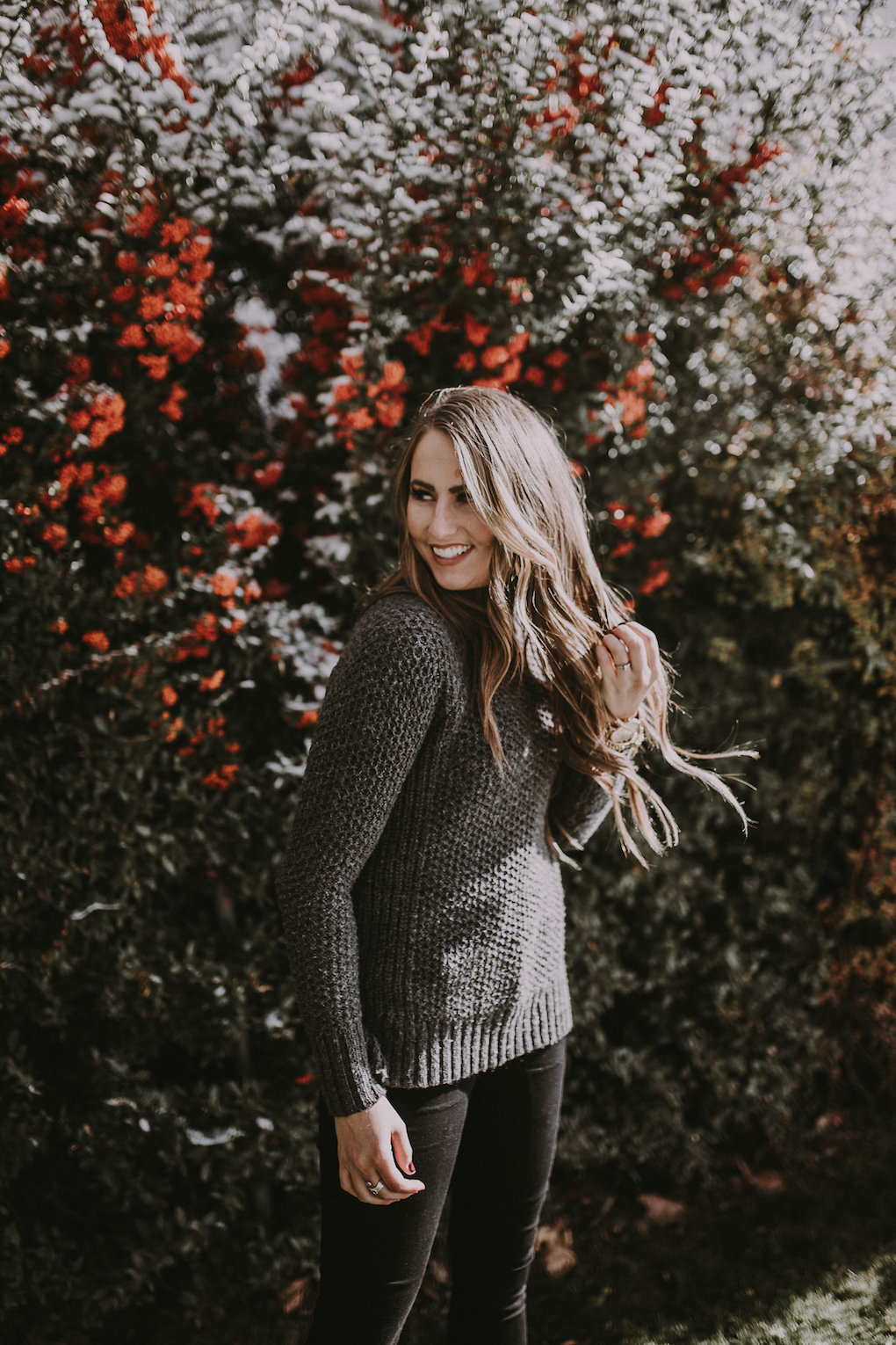 girl standing in front of berry bush wearing a thick oversized grey knit turtleneck with black jeans and black booties with brown loosely curled hair