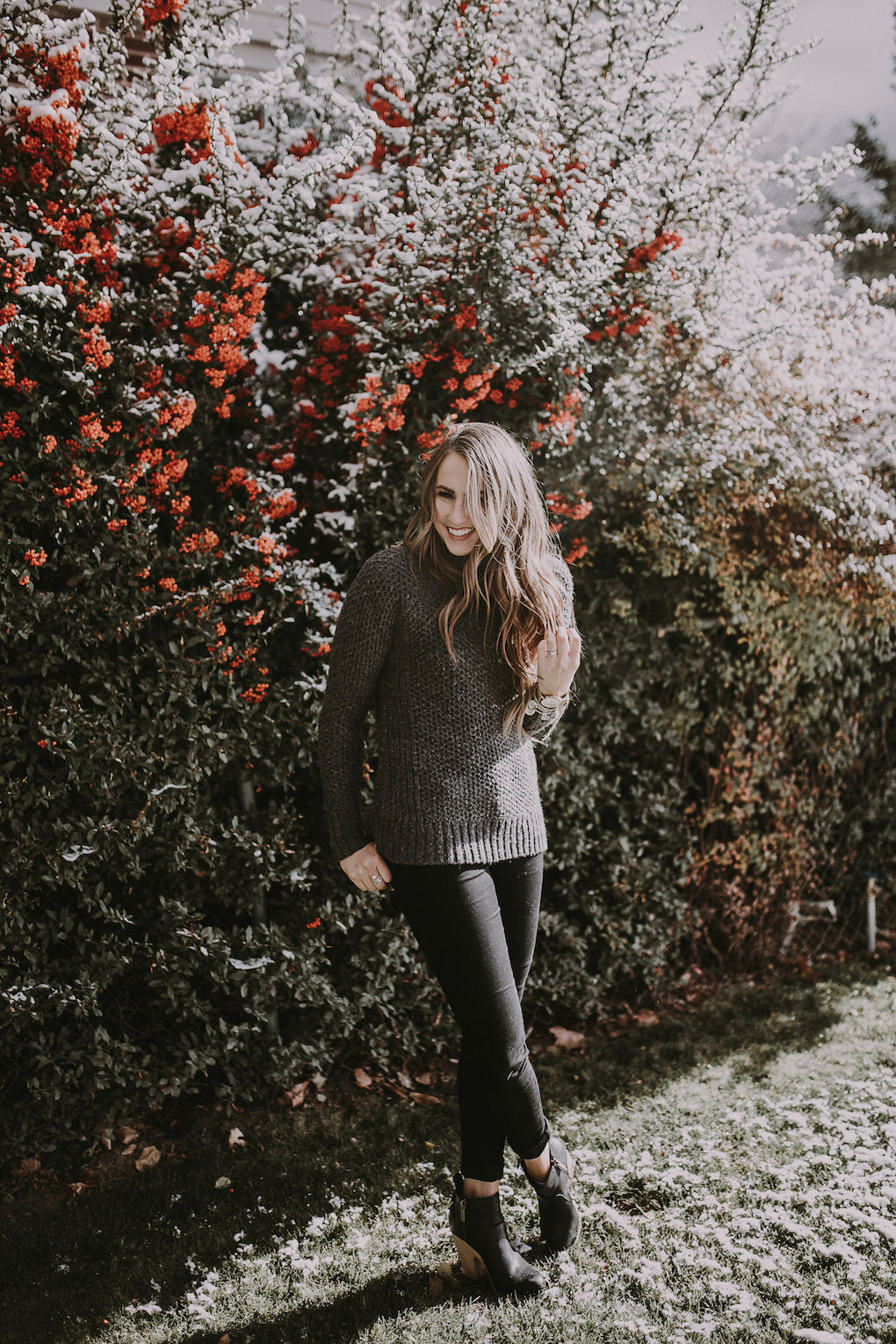 girl standing in front of berry bush wearing a thick oversized grey knit turtleneck with black jeans and black booties with brown loosely curled hair