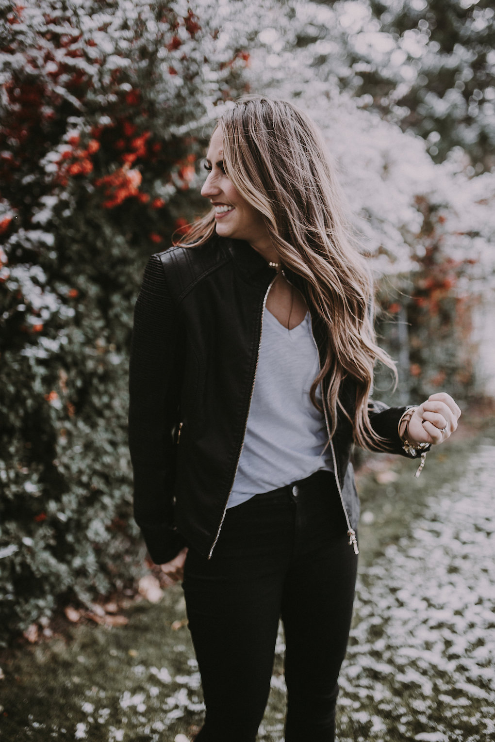 girl standing in front of berry bush wearing a black leather jacket and basic white tee paired with black jeans with holes in the knees and blcak booties with brown loosely curled hair