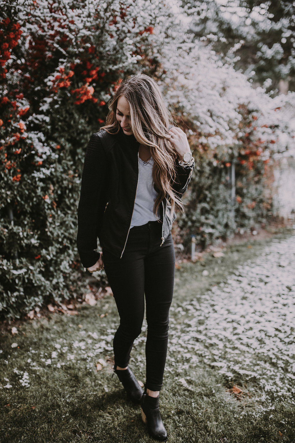 girl standing in front of berry bush wearing a black leather jacket and basic white tee paired with black jeans with holes in the knees and blcak booties with brown loosely curled hair