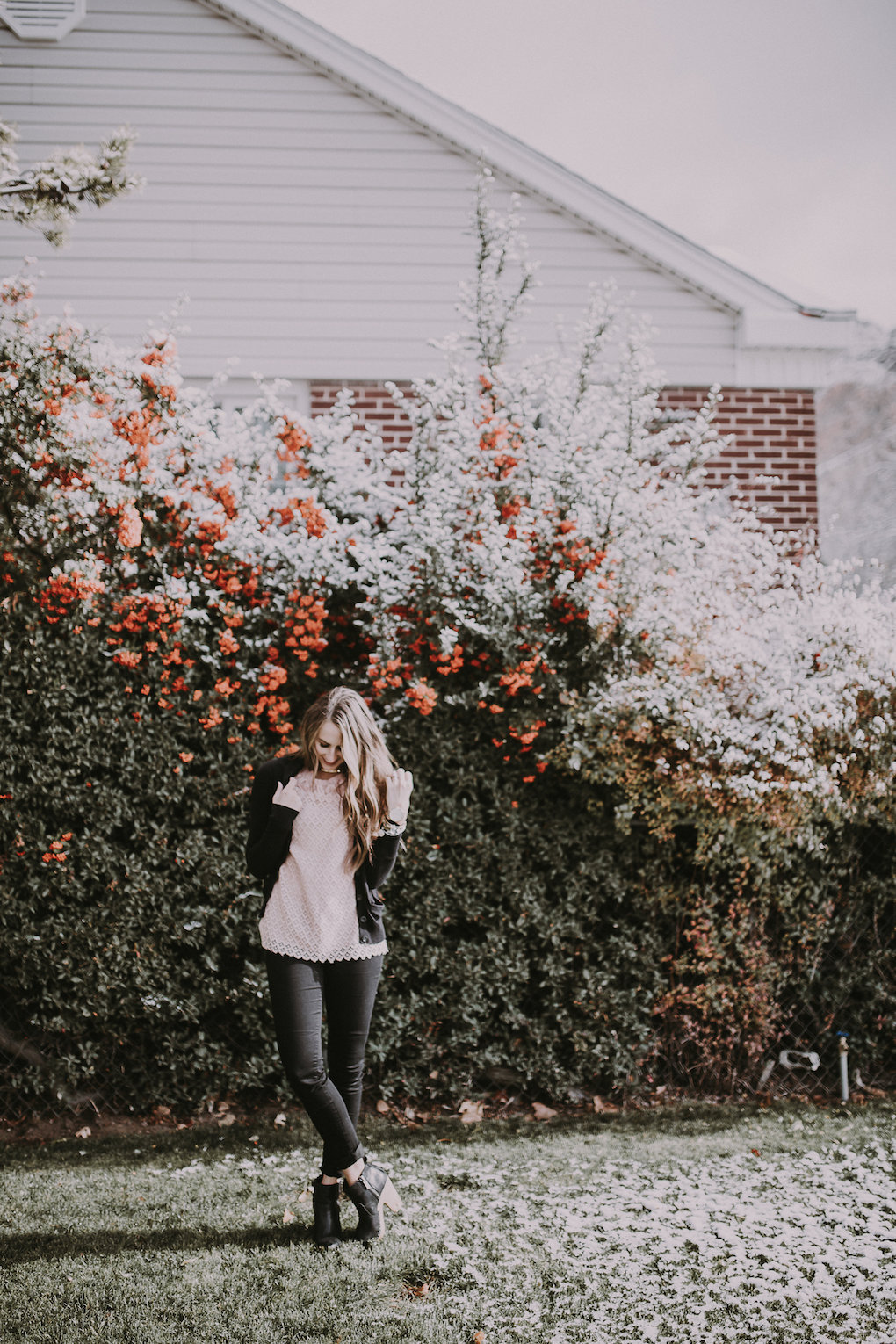 girl standing in front of berry bush wearing a light pink lace top with a black cardigan and black distressed jeans and blck booties with brown loosely curled hair