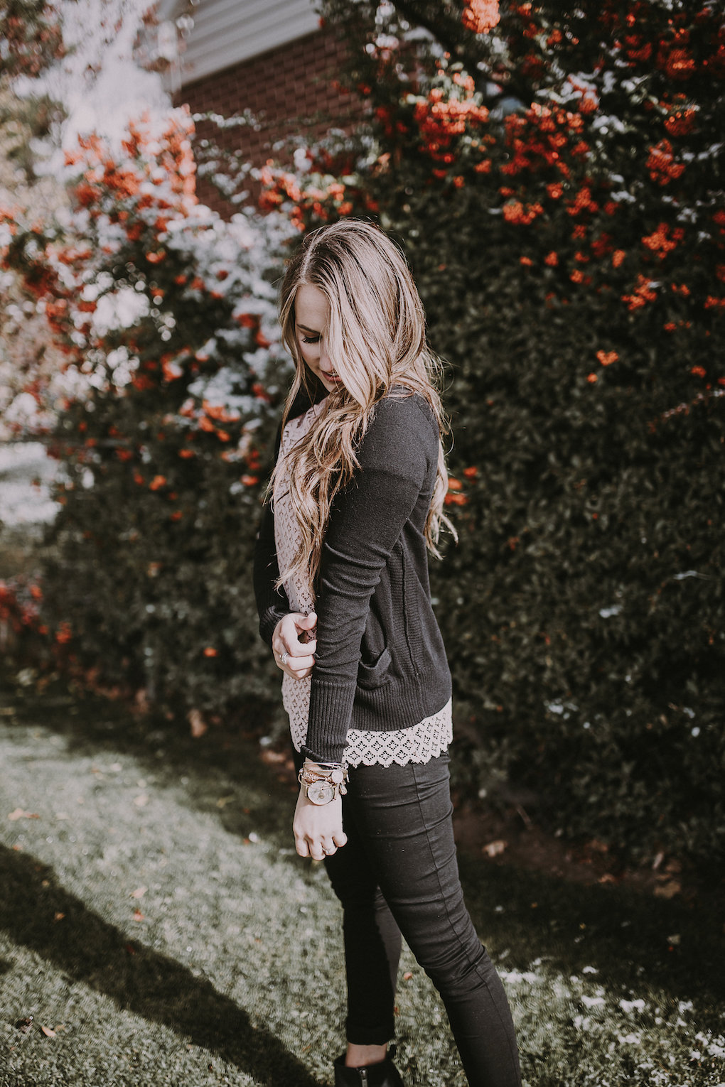 girl standing in front of berry bush wearing a light pink lace top with a black cardigan and black distressed jeans and blck booties with brown loosely curled hair